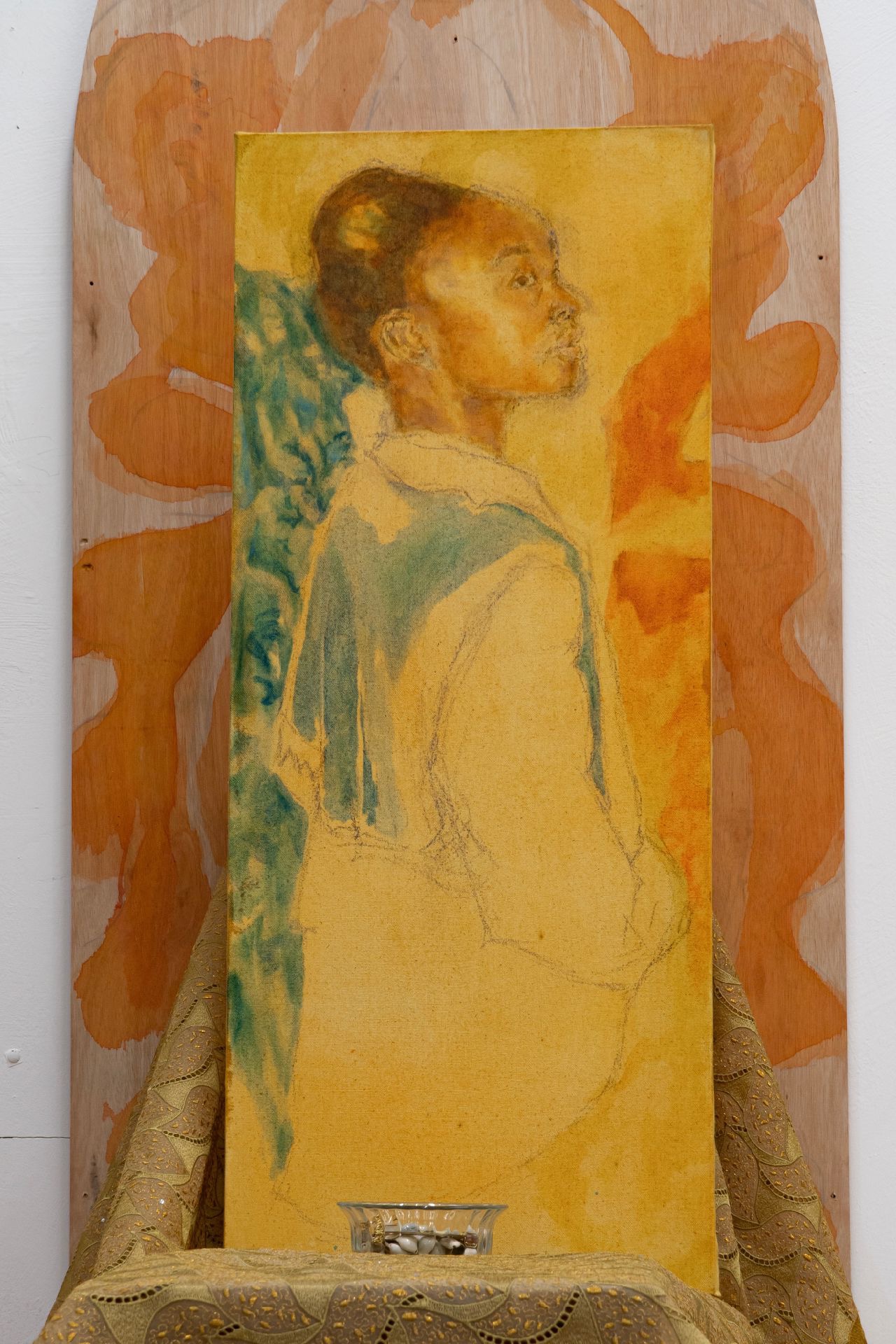 This is a detail of a painting of a young girl looking away from the viewer, the predominant colours in the work are yellow, blue and red. The painting is from an installation work, she is resting on gold/yellow lace fabric from Nigeria, at the front of th