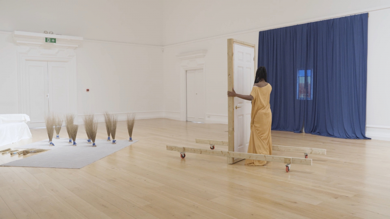 Installation view showing a woman dressed in yellow in front of a free-standing door. A blue curtain drapes one wall with a small projection in the middle, and dried grasses are displayed in bunches to the left.